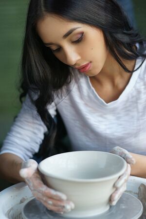 Young beautiful pretty woman with brunette dark hair working on pottery wheel and sculpting clay potの写真素材