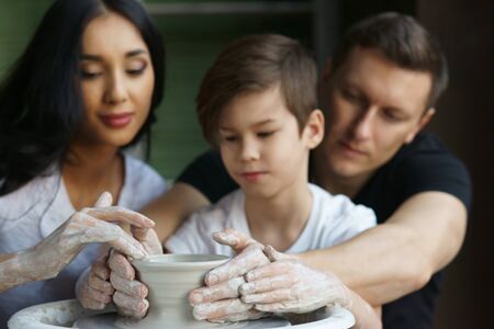 Family working on pottery wheel. Handsome man, beautiful pretty brunette woman and son. Shallow DOF. Focus on hands on pot clay.の写真素材