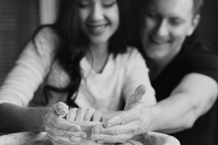 Pottery. Happy joyous romantic caucasian couple working together on potters wheel and sculpting or making clay pot. Handsome man and beautiful pretty brunette woman. Focus on dirty hands.の写真素材
