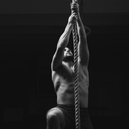 Male athlete or sportsman climbing rope at dark gym. Topless handsome muscular man in gray shorts.の写真素材