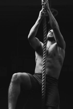 Male athlete or sportsman climbing rope at dark gym. Topless handsome muscular man in gray shorts.