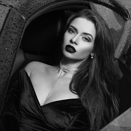 Fashion portrait of young beautiful woman with red lips and long straight brunette hair wearing brilliant shining earrings, necklace choker and blue evening dress sitting in red luxury sport carの写真素材