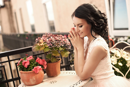 Outdoor portrait of young beautiful pretty caucasian brunette bride posing in white fluffy dress. Attractive girl sitting at terrace on summer sunny day.の写真素材
