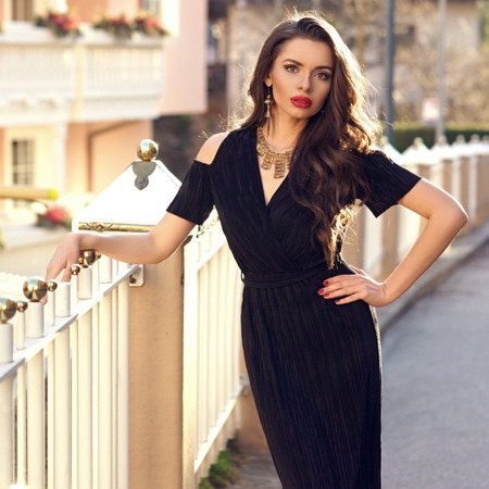 Gorgeous female model with red lips, long wavy hair and luxury golden jewelry, dressed in black dress with cut-out shoulders, posing against white railing and beautiful building on background.の写真素材