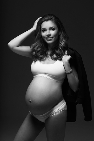 Full body portrait of attractive smiling pregnant brunette woman with slightly curly hair, wearing white sports underwear, trainers and with black jacket draped over shoulders, standing and posing.の写真素材
