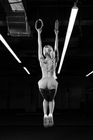 Slim beautiful female gymnast with long blonde braid, dressed in pink crop top, shorts and white trainers, hanging on rings in gym. Gorgeous young woman performing gymnastics exercise. Back view.の写真素材