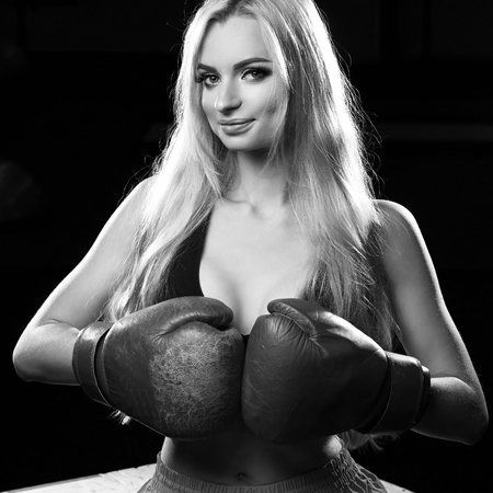 Gorgeous smiling blonde woman with long hair, dressed in black fitness bra, pink shorts and red boxing gloves, sitting on wooden box with her arms touching each other. Beautiful female boxer posing.の写真素材