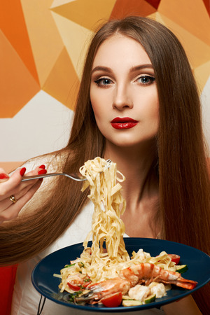 Portrait of attractive long haired woman with slightly ajar mouth and red lips eating Italian pasta with seafood and vegetables. Gorgeous young female model posing with delicious fettuccine on fork.の写真素材