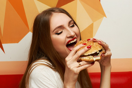 Portrait of gorgeous young woman with closed eyes and red lips holding tasty cheeseburger and biting it. Attractive female model eating burger with pleasure. Beautiful girl enjoying fast food meal.の写真素材