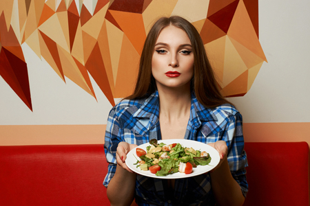 Portrait of seductive young woman dressed in checkered unbuttoned shirt holding plate with delicious vegetable salad made of shrimps, tomatoes, avocado. Sexy female model demonstrating tasty meal.の写真素材