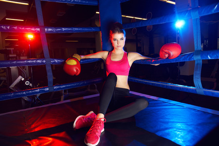 Full body portrait of boxer woman with long dark hair pulled back in pony tail, wearing pink sports bra, leggings, trainers and red boxing gloves, sitting in corner of ring and leaning on ropes.の写真素材