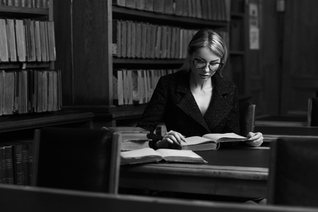 Beautiful blonde woman wearing elegant black tweed jacket and glasses sitting at desk beside bookshelf and reading book. Young gorgeous female student studying at library. Smart is new sexy concept.の写真素材