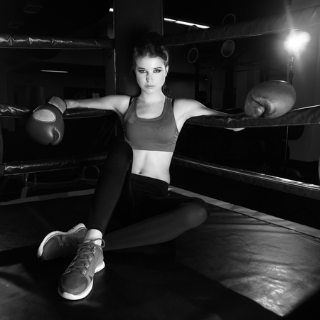 Full body portrait of boxer woman with long dark hair pulled back in pony tail, wearing pink sports bra, leggings, trainers and red boxing gloves, sitting in corner of ring and leaning on ropes.の写真素材