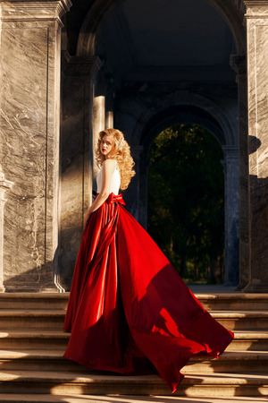 Fashion style photo of young beautiful woman in evening dress with white top and red flying skirt posing at stairs at sunsetの写真素材