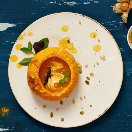 Delicious vegetable soup decorated with basil leaves and cooked in pumpkin lying on flat round plate standing on blue aged wooden surface. Tasty restaurant vegetarian meal, veggie recipe. Top view.の写真素材