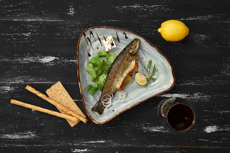 Fried fish served with lemon slices, lettuce, tomatoes, rosemary and onion rings lying on plate, glass of red wine, whole lemon, bread sticks and crispbread on blue old wooden surface. Top view.の写真素材