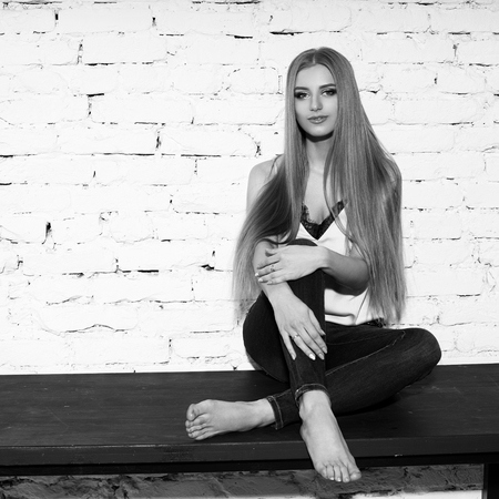 Young beautiful woman with long straight blonde hair in blue jeans and bright blouse sitting on black wooden table with crossed legs and looking in camera.の写真素材