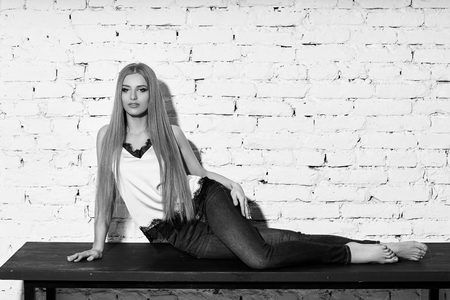 Young beautiful woman with long straight blonde hair in blue jeans and bright blouse sitting on black wooden table with crossed legs and looking in camera.の写真素材