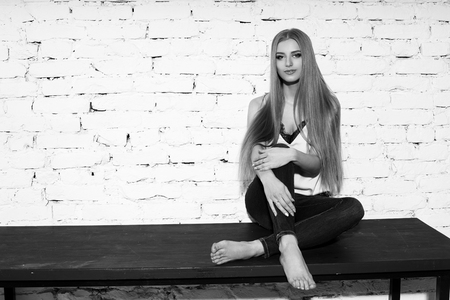 Young beautiful woman with long straight blonde hair in blue jeans and bright blouse sitting on black wooden table with crossed legs and looking in camera.の写真素材