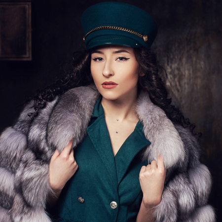 Stylish woman wearing green jacket dress and cap, fur coat and black jackboots posing in dark interior. Fashion style portrait. Brunette woman with curly hair.の写真素材
