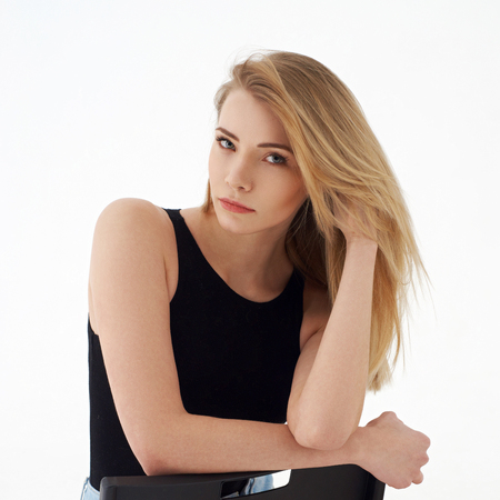 Young beautiful sexy woman in blue jeans shorts and black shirt posing and sitting on simple black chait in white studio. Fashion model with long straight hair.の写真素材