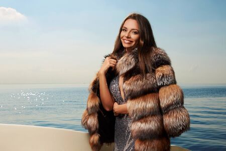 Young fashionable gorgeous glamorous woman in elegant silver shining evening dress and striped fur coat with long straight brown hair standing and posing at luxury yacht on a sunny summer dayの写真素材