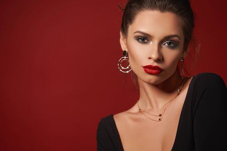 Closeup fashion vogue style portrait of young elegant woman with hairstyle and makeup. Girl with red lips wearing earrings and necklace. Studio shot against red backgroundの写真素材
