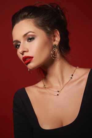 Closeup fashion vogue style portrait of young elegant woman with hairstyle and makeup. Girl with red lips wearing earrings and necklace. Studio shot against red backgroundの写真素材