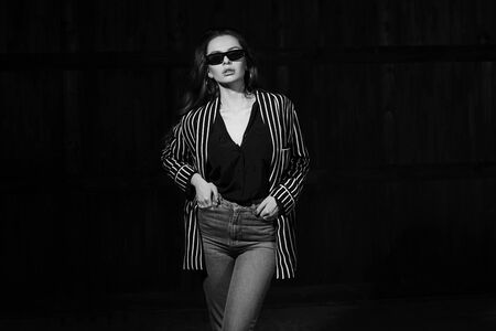 Body length interior portrait of stylish young female modelwearing grey jeans and striped blazer standing and posing in dark room. Vogue style. Studio shotの写真素材