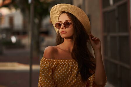 Closeup outdoor portrait of young beautiful caucasian young woman with long brunette hair wearing yellow polka dot dress, sunglasses and thatch hat. Summer sunny evening streetの写真素材