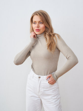 Woman with long blonde hair wearing a beige turtleneck and white high-waisted pants poses with one hand on her face and the other in her pocket, looking contemplativeの写真素材