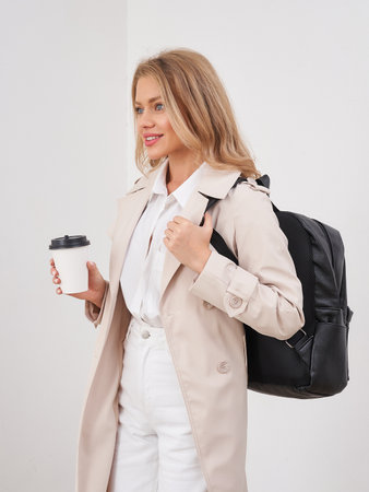 Smiling woman with long blonde hair wearing a beige coat over a white shirt. She holds a coffee cup...
