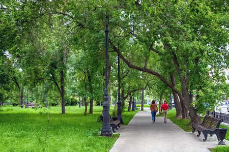 Two girls are walking in a green city park with empty benches, lampposts and lush deciduous trees. Summer. Day.の写真素材