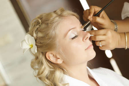 Beautiful, cute blond bride doing makeup before wedding day. Long lashes, white flowers in hair, smilling and happy.の写真素材