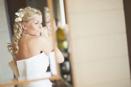 Gorgerous bride standing in front of mirror and fixing her hair and make up. Putting earings, powdering and smilling.の写真素材