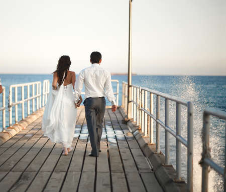 Gorgeous wedding couple walking through pier, splashes and drops on sunset. Beautiful wooden old pier pacific or atlantic ocean.の写真素材