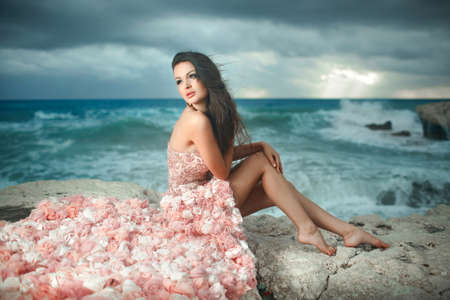 Beautiful, charming and sexy woman, model sitting on rocks, cliffs during sunset. Windy day at paradise.の写真素材