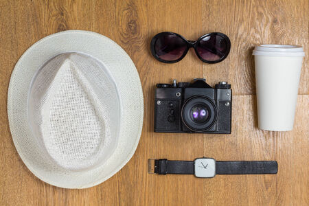  Travel concept. Top view of braided hat, pair of glasses, vintage camera, takeaway cup, watch and cute cloth heart on a wooden background.の写真素材