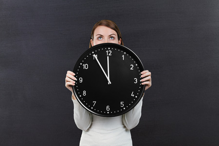 Woman holding clock over her face on chalkboard background with copy spaceの写真素材