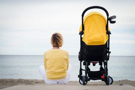 Woman looking through the sea while sitting on beach with baby carriage. Young mother sitting outdoor with stroller. Maternity concept with copyspaceの写真素材
