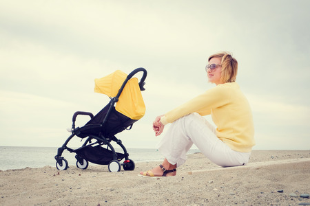 Smiling mother sitting on beach by the sea next sleeping baby in strollerの写真素材