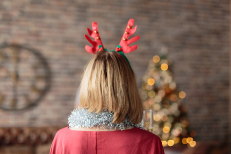 Woman wearing antler deer headband looking at decorated Christmas tree. Rear view. Christmas tree and wall clock on background. Christmas conceptの写真素材
