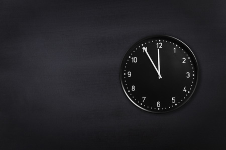 Black wall clock showing five minutes to midnight on black chalkboard background. Office clock showing 5 min to midnight or midday on black textureの写真素材