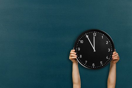 Man hold round wall clock on chalkboard background. Male hands holding round black clock showing 5 minutes to midnightの写真素材