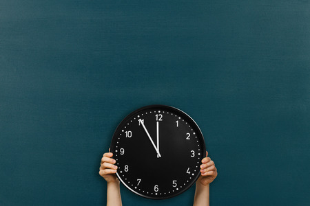 Man holds round office clock showing 5 minutes to midday on chalkboard background. Crop imageの写真素材