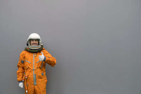Happy astronaut wearing orange space suit and helmet showing thumbs up gesture on a gray wall backgroundの写真素材