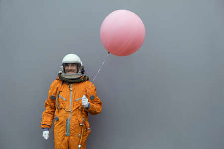 Astronaut wearing orange space suit and helmet holding pink inflatable balloon against gray wall backgroundの写真素材