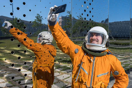 Happy astronaut wearing orange space suit and space helmet holding mobile phone and taking selfie outdoors.の写真素材
