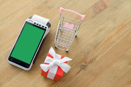 POS terminal with blank screen with miniature shopping cart and red gift box on a wooden backgroundの写真素材