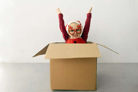 A child wearing funny Christmas glasses and Santa hat raised her hands up while sitting in a cardboard box against white wall backgroundの写真素材